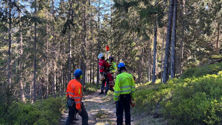 Zwei Rettungskräfte von DRF Luftrettung und Bergwacht hängen kurz über dem Boden schwebend in der Seilwinde, sie vom Rettungshubschrauber kommt.