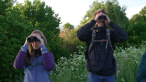 Eine Frau und ein Mann stehen in Oudoor-Klamotten und mit einem Rucksack auf dem Rücken nebeneinander. Sie halten sich beide ein Fernglas vor die Augen.