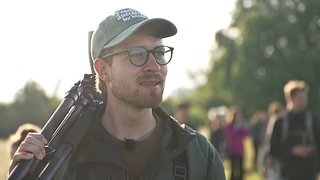 Ein junger Mann mit Brille, Cappy und einem Stativ über die Schulter getragen schaut in die Ferne.