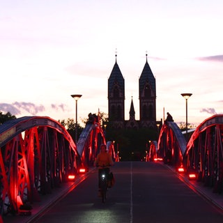 Die Wiwili-Brücke in Freiburg, auch Blaue Brücke genannt, erstrahlt an diesem Abend in rotem Licht. Eine Frau fährt auf einem Fahrrad über die Brücke. 