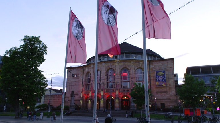 Rote Fahnen mit SC Freiburg-Logo wehen auf einem Platz in Freiburg. Im Hintergrund ist das rot beleuchtete Stadttheater zu sehen. 