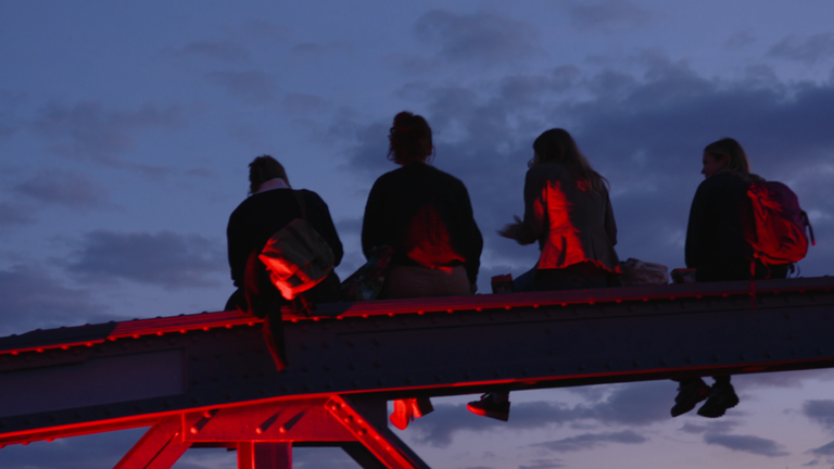Vier Frauen sitzen abends auf einer rot beleuchteten Brücke in Freiburg.