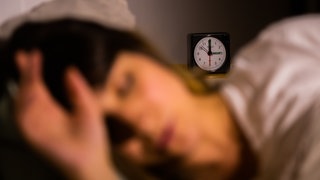 Eine Frau liegt im Bett mit einem Wecker im Hintergrund. Eine Studie empfiehlt bei Schlafstörungen, gezielt weniger zu schlafen.