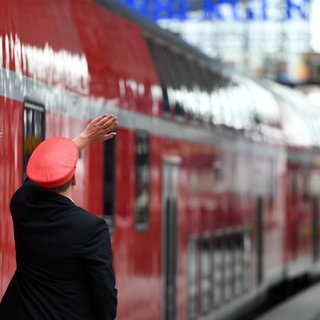 Ein Bahnmitarbeiter gibt an einem Bahnsteig ein Handsignal zur Weiterfahrt. 