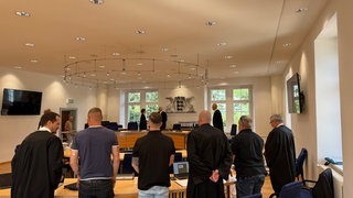 In einem Gerichtssaal stehen sechs Männer im Vordergrund. 