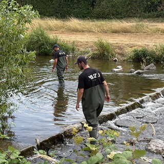 Zwei Männer mit Cappy  und schwarzen Tshirts und der Aufschrift "Polizei" steigen in einen kleinen Fluss.