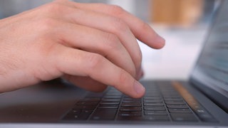 Symbolbild: Seitenansicht einer Hand an einem Laptop. Die Person tippt auf die Tastatur.
