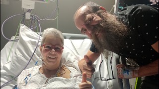 Eine Frau liegt in einem Krankenhausbett, daneben steht ein Mann mit Vollbart. Beide strecken ihre Daumen nach oben. 