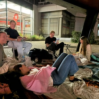 Junge Leute campieren mit Decken im Freien auf dem Platz vor einem Sparkassengebäude.