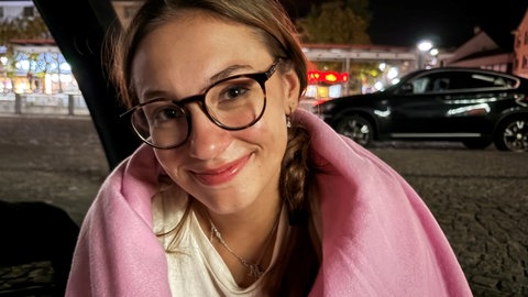 Junge Frau mit Brille, eingewickelt in eine Decke, vor nächtlichen Lichtern der Stadt.