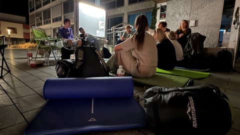 Junge Leute mit Isomatte und Schlafsack auf einem Platz unter dem Vordach eines Gebäudes.