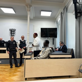 Ein Mann steht vor dem Landgericht Ravensburg wegen Mordes durch Messerstiche vor einem Supermarkt in Ravensburg. Die Frau neben ihm (li.) ist eine Dolmetscherin.