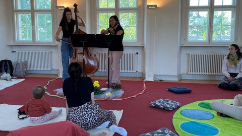 Babys mit Eltern oder Großeltern sitzen und liegen auf Teppichen und hören sich ein Konzert mit klassischer Musik an. 
