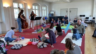 Babys mit Eltern oder Großeltern sitzen und liegen auf Teppichen und hören sich ein Konzert mit klassischer Musik an. 