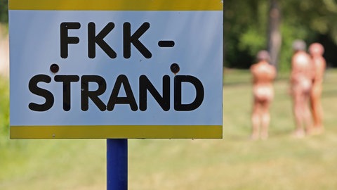FKK-Strand Schild mit drei Menschen im Hintergrund