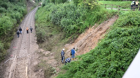 Erde liegt nach einem Hangrutsch auf den Gleisen, im Hintergrund ist der entgleiste Regionalexpress auf der Strecke zu sehen. Bei dem Zugunglück im Südosten Baden-Württembergs sind drei Menschen ums Leben gekommen.