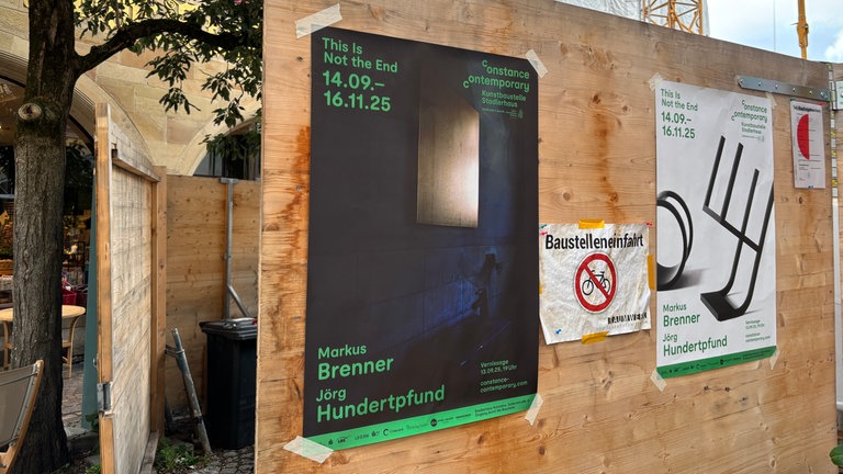 Ein Bauzaun mit Ausstellungsplakaten. Im 2024 ausgebrannten Stadlerhaus in Konstanz ist jetzt eine Doppelausstellung zu sehen.