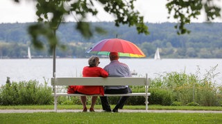 Ein Mann und eine Frau sitzen am Ufer des Bodensee mit einem Regenschirm  in der Hand auf einer Bank.