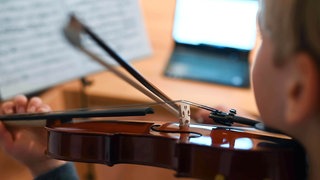 Ein Mann spielt Geige vor seinem Laptop. Die Bodensee-Ouvertüre ist das erste Werk der Bodensee Philharmonie, das von einer KI komponiert worden ist. (Symbolbild)