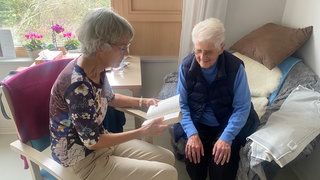 Zwei Frauen sitzen zusammen und lesen ein Buch. Medienbotin Christina Waldschütz ist mit einem Buch zu Besuch bei Maria Gottstein.