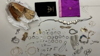 Ein aufgebrochener Brotlaib und viele darin versteckte Schmuckstücke sind auf einem Tisch ausgebreitet.