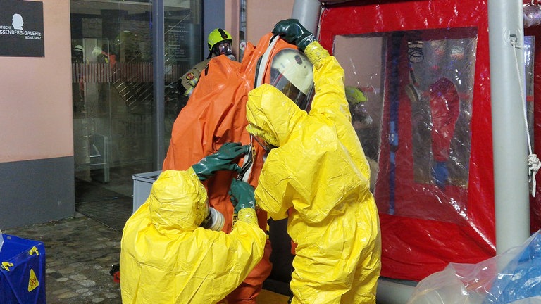 Zwei Einsatzkräfte helfen einem Feuerwehrmann dabei, einen Schutzanzug nach dem Gefahrstoffeinsatz in Konstanz auszuziehen.