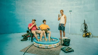 Szenenbild aus der "Tschappel"-Folge "Oben ohne Down Under": In einer blau angestrichenen Güllegrube sitzen BlaBla (Sebastian Jakob Doppelbauer) und Carlo (Jeremias Meyer) nebeneinander auf Stühlen. Sie haben ihre Füße in einen aufblasbaren Kinderpool gestellt. Aydin (David Ali Rashed) steht daneben. Mit Dosenbier in der Hand genießen sie ihre Pause. Neben dem Pool liegen Malerutensilien und ein Hochdruckreiniger.