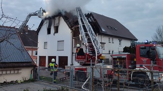 Rauch steigt auf von einem brennenden Haus, im Vordergrund die Feuerwehr. Nach dem Brand ist das Wohnhaus in Stockach vorerst nicht bewohnbar.