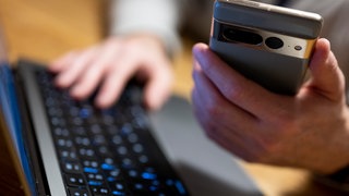 Ein Mann sitzt vor einem Laptop, die eine Hand auf der Tastatur. Mit der anderen Hand bedient er ein Smartphone.