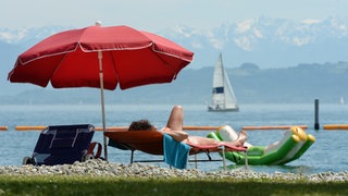 Ein Badegast liegt am Ufer des Bodensees auf einer Liege unter einem Sonnenschirm. An Pfingsten erwarten Hotels und Campingplätze einen großen Urlauberansturm.