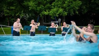 Musikerinnen und Musiker sind an ungewöhnlichen Orten zu sehen: Im Wasserbecken, im Riesenrad, unter einer Brücke.