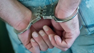 Zwei Hände, die in Handschellen hinter dem Rücken gefesselt sind. (Symbolbild)