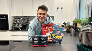 Der Influencer Sven Teichmann steht in seiner Küche und hält seine zwei Bücher in der Hand. Er ist selbstständig.