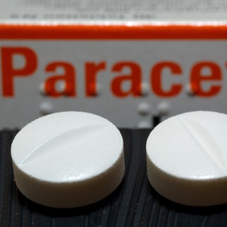 Vor einer Arzneiverpackung liegen Tabletten, die den Wirkstoff Paracetamol enthalten.