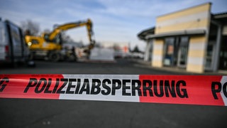 Ein Bagger steht nach einer Zerstörungsfahrt neben einem Autohaus in Tauberbischofsheim hinter einer Polizeiabsperrung (Archivbild). Jetzt richten sich die Ermittlungen der Staatsanwaltschaft gegen fünf Polizeibeamte, die die tödlichen Schüsse gegen einen Baggerfahrer abgegeben haben sollen. 