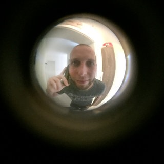 Ein Mann mit Handy am Ohr schaut durch einen Türspion (Symbolbld). Die Polizei warnt vor Trickdieben, die immer häufiger an der Haustür klingeln. 