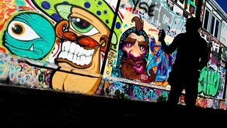 Ein Sprayer steht mit einer Spraydose vor einer Wand mit zahlreichen Graffitis (Symbolbild). 