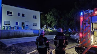 Zwei Feuerwehrleute stehen neben einem Löschfahrzeug, vor einem neu gebauten Haus. In der Nacht hat es in dem Haus in Künzelsau gebrannt. Die Nachbarn haben die Bewohner über Leitern gerettet.