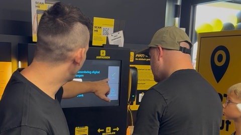 Ein Mitarbeiter des ORTkauf erklärt einem Kunden, wie die Selbstebdienungskasse funktioniert. In Weißbach hat ein personalloser Mini-Supermarkt eröffnet. Der Shop soll sieben Tage die Woche, rund um die Uhr geöffnet haben. Betreiber ist ein Start-up aus Schwäbisch Hall.