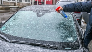 Ein Mann sprüht Einteiserspray auf eine zugefrorene Frontscheibe. (Symbolbild)