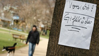 (Symbolbild) Ein Schild mit der Aufschrift "Vorsicht Giftköder" hängt im Vordergrund an einem Baum. Im Hintergrund ist unscharf eine Person mit einem Hund an der Leine zu sehen. - Vergangene Woche sind in einem Waldstück zwischen Obersulm-Eschenau (Kreis Heilbronn) und Bretzfeld-Waldbach (Hohenlohekreis) zwei Hunde vermutlich an Giftködern gestorben. (Symbolbild)