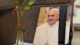 Bild des verstorbenen Papst Franziskus in einer Kirche, davor eine Kerze und Blumen (Archivbild).