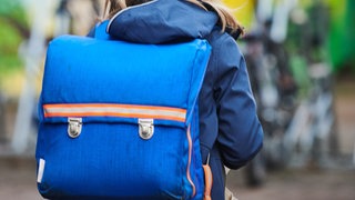 Ein Kind mit langen Haaren und einem blauen Rucksack, von hinten zu sehen