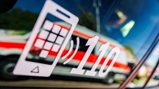 Die Nummer des Polizeinotrufs 110 steht auf der Scheibe eines Polizeifahrzeugs, in der sich ein Fahrzeug vom Roten Kreuz spiegelt. (Symbolbild)
