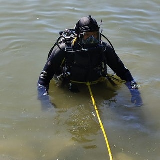 Taucher im trüben Wasser eines Sees, ein gelbes Seil ist um seinen Körper gebunden und geht zum Ufer.