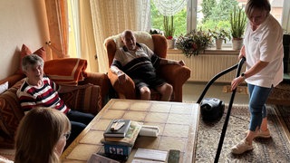 Eine Haushaltshilfe saugt in einem Wohnzimmer den Teppich, während ein älterer Mann auf einem Sessel sitzt, eine ältere Frau auf einem Sofa und eine jüngere Frau sich Briefe anschaut.