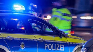 Ein Streifenwagen der Polizei bei Nacht, im Hintergrund ist unscharf ein Beamter in einer gelben Warnjacke zu erkennen. (symbolbild): Auf der Flucht vor der Polizei hat ein 27-Jähriger bei Schwaigern (Kreis Heilbronn) einen tödlichen Unfall verursacht. Sein 20-jähriger Beifahrer starb.