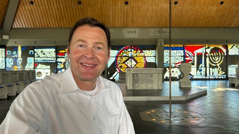 Der Pfarrer der Autobahnkirche Baden-Baden sitzt auf einer Bank in der Kirche. Die Kirche steht auf einem Rasthof für Reisende der A5.