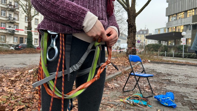 Eine Person zieht sich einen Klettergurt an. Sie gehört zu der Gruppe, die die Platanen vor dem Badischen Staatstheater in Karlsruhe besetzen.