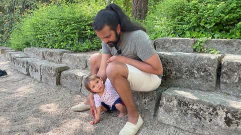 Vater sitz mit kleiner Tochter auf einer Treppe am Spielplatz: Benedict sieht es für sich als Vater als Chance, dass es keinen Mutterinstinkt gibt. Er teilt sich die Elternschaft mit seiner Frau gleichberechtigt.